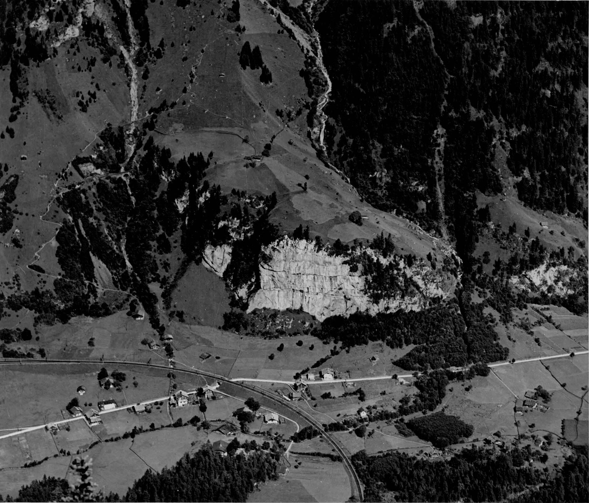 Historische Schwarzweissaufnahme um 1930 vom Kirchhorn gegen Osten: helle Felswand der Fluh über dem Talboden von Mitholz, davor Bahnkurve, Strasse und verstreute Häuser.