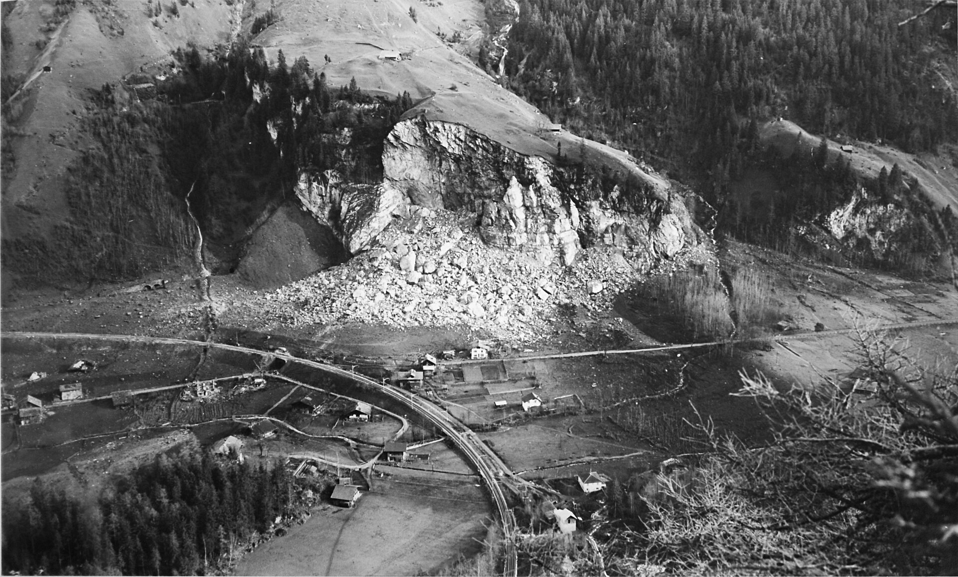 Schwarzweissaufnahme vom Januar 1948 von Westen: die ausgebrochene Felswand der Fluh bei Mitholz mit grossem hellem Schuttkegel, darunter die Bahnkurve der BLS und verstreute Häuser im Talboden.