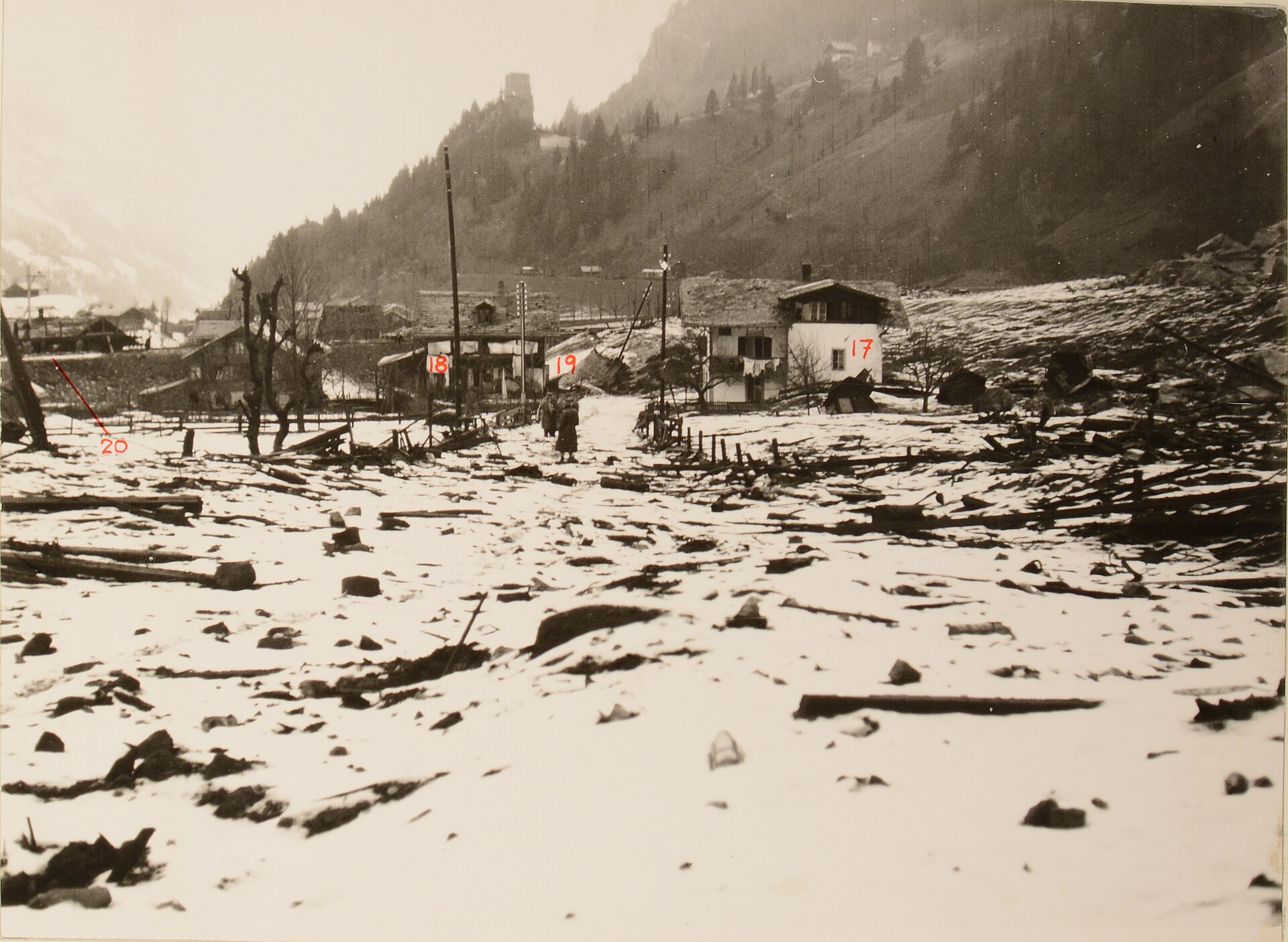 Polizeifoto vom 26. Dezember 1947: verschneite, verschüttete Staatsstrasse bei Mitholz, übersät mit Steinblöcken und Balken, dahinter beschädigte Häuser mit Nummernmarkierungen.