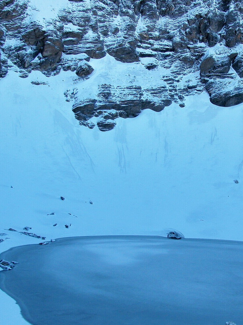 Hochformat-Ausschnitt: zugefrorene Fläche des Roopkund-Sees vor verschneiten Felswänden
