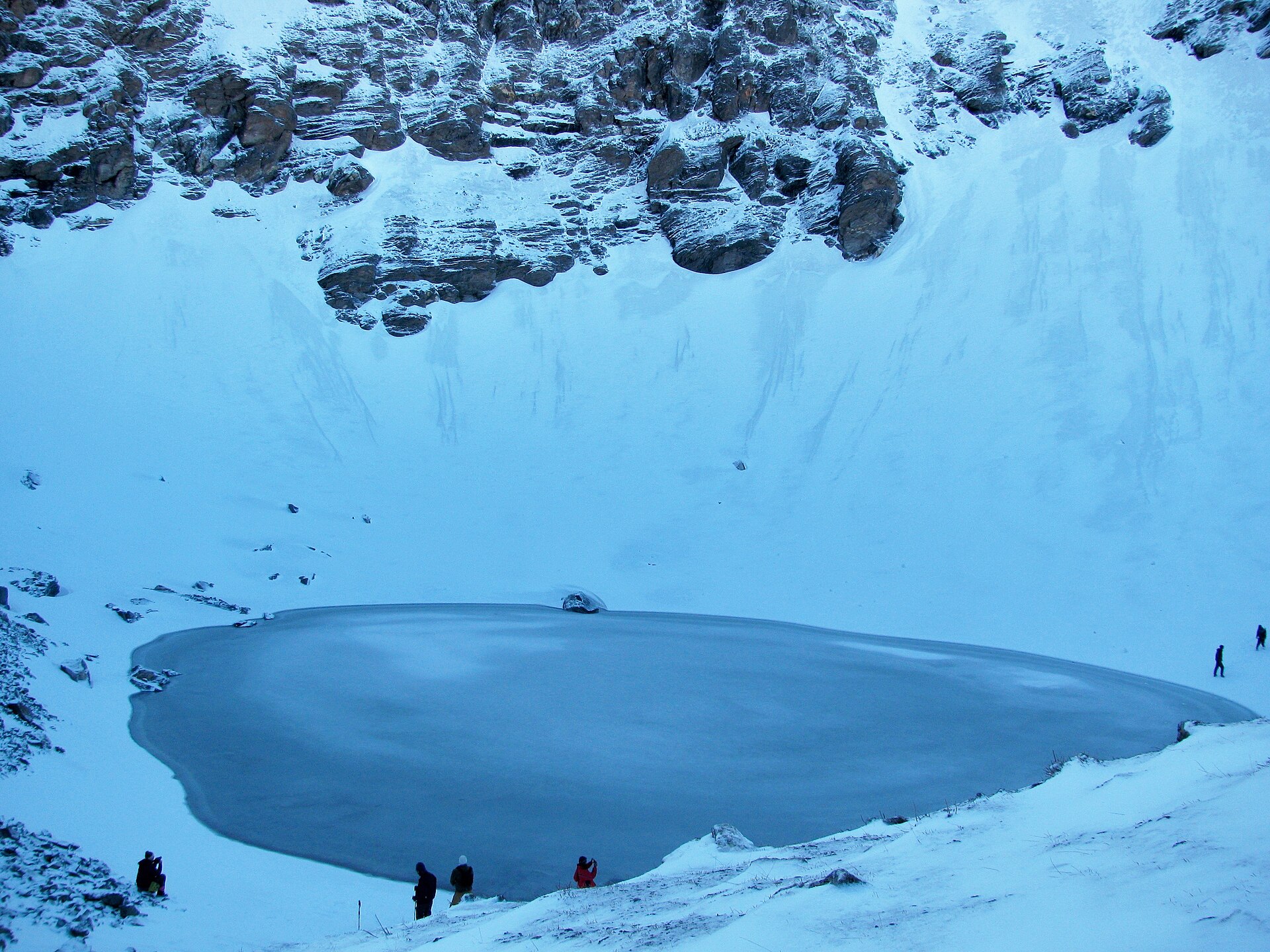 Zugefrorener Roopkund-See unter steilen Felswänden, am Ufer eine kleine, nicht erkennbare Wandergruppe